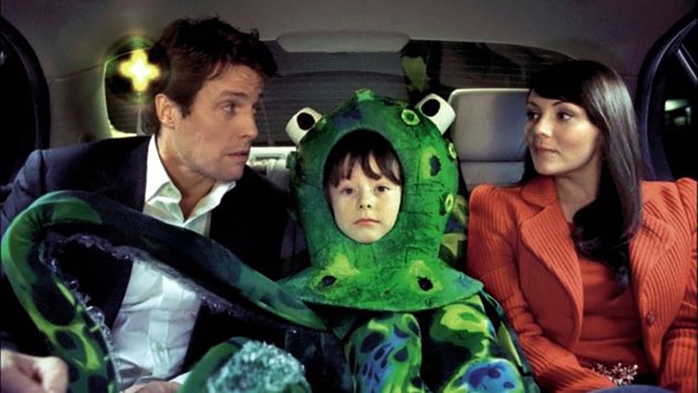A man in a suit, a child dressed in a green octopus costume, and a woman in a red jacket sit together in the back seat of a car, looking at each other.