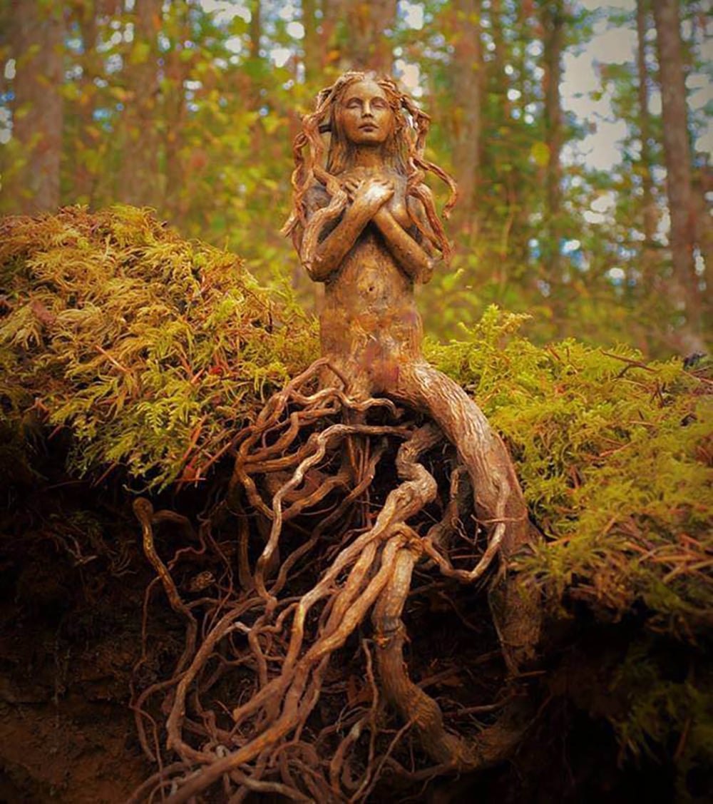 A wooden sculpture resembling a woman with crossed arms, blending into tree roots at her base, stands among green moss and forest surroundings.