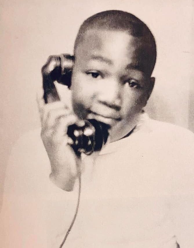A young boy in a white shirt holds an old-fashioned telephone receiver to his ear, looking slightly to the side with a gentle expression. The photo is in black and white.
