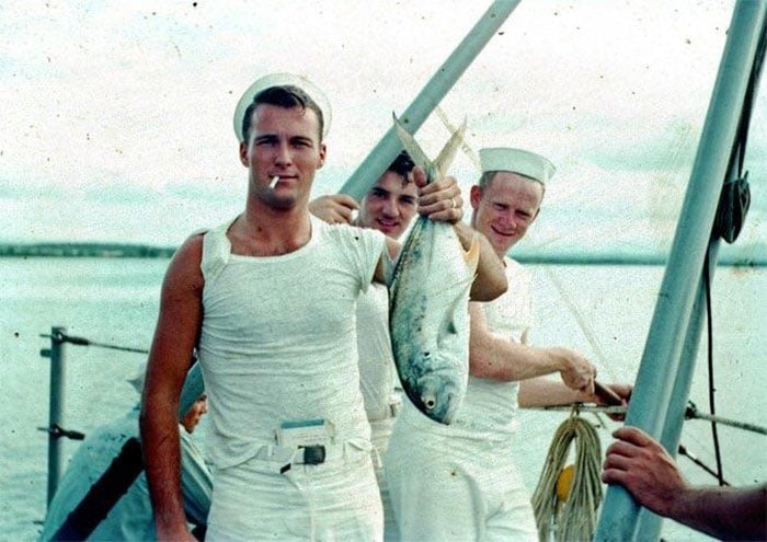Three young sailors in white uniforms stand on a boat. One sailor in front, smoking a cigarette, proudly holds up a large fish. The other two sailors smile in the background, posing with ropes and equipment. Water is visible behind them.