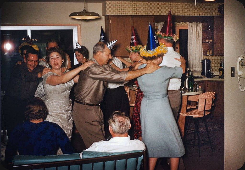 A group of adults wearing party hats dance in a conga line at a lively indoor party, with festive decorations and drinks visible in a cozy, mid-century kitchen and living room.