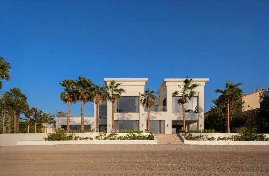 Modern beachfront villa with large glass windows, white exterior, and two main sections, surrounded by palm trees and greenery under a clear blue sky. Sandy area is visible in the foreground.