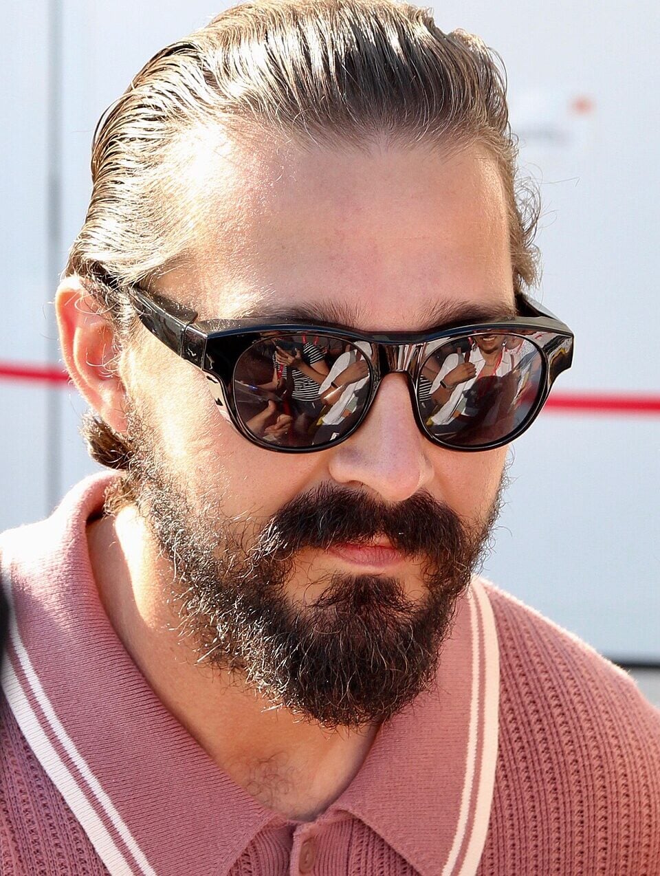 A man with slicked-back hair, a full beard, and mustache, wearing dark sunglasses and a pink collared shirt, stands outdoors in bright light. The sunglasses reflect people and surroundings.