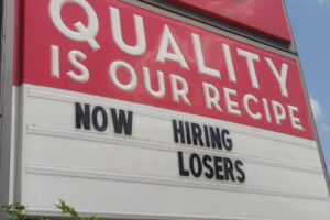 A Wendy's restaurant sign reads "QUALITY IS OUR RECIPE" and below it, the marquee says "NOW HIRING LOSERS." The sky is blue with scattered clouds, and shops are visible in the background.