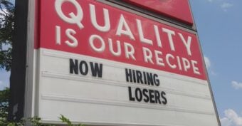 A Wendy's restaurant sign reads "QUALITY IS OUR RECIPE" and below it, the marquee says "NOW HIRING LOSERS." The sky is blue with scattered clouds, and shops are visible in the background.