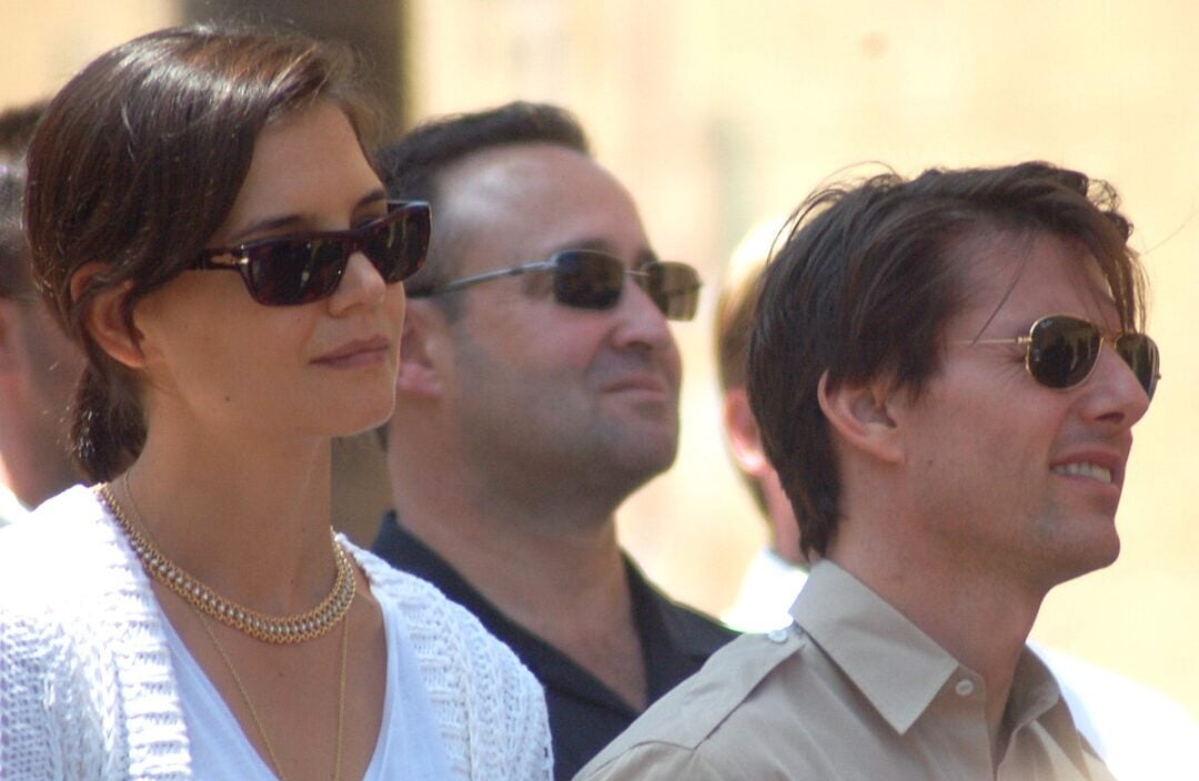 A woman and a man wearing sunglasses stand outdoors among a group of people. The woman has short dark hair and wears a white sweater and gold necklace; the man has short brown hair and wears a light shirt.