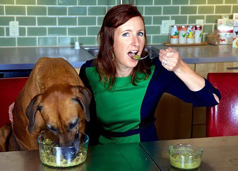 A woman in a green dress eats from a spoon while sitting next to a large brown dog, who is eating from a bowl on the table. They both appear to be sharing a meal in a modern kitchen with a green tiled backsplash.
