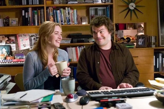 A woman smiles while holding a mug and sitting next to a man at a desk cluttered with office supplies and a computer keyboard; bookshelves and a clock are visible in the background.