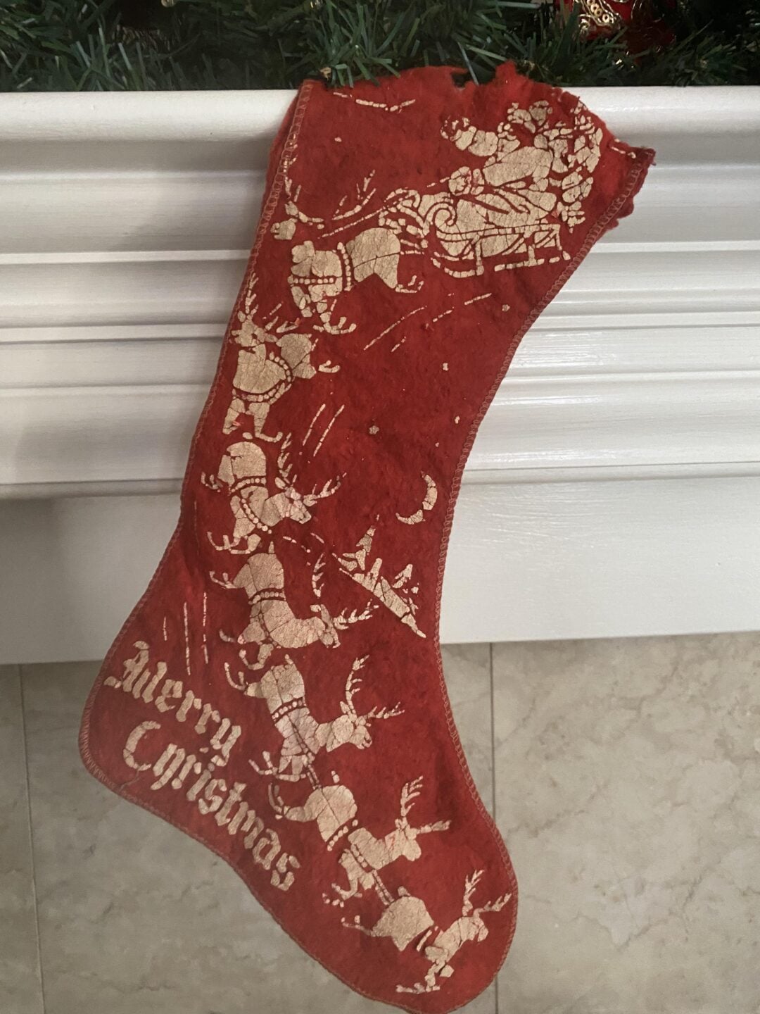 A red Christmas stocking with a gold design of Santa Claus in his sleigh pulled by reindeer, flying over rooftops, and the words “Merry Christmas” at the bottom. The stocking hangs from a white mantle.