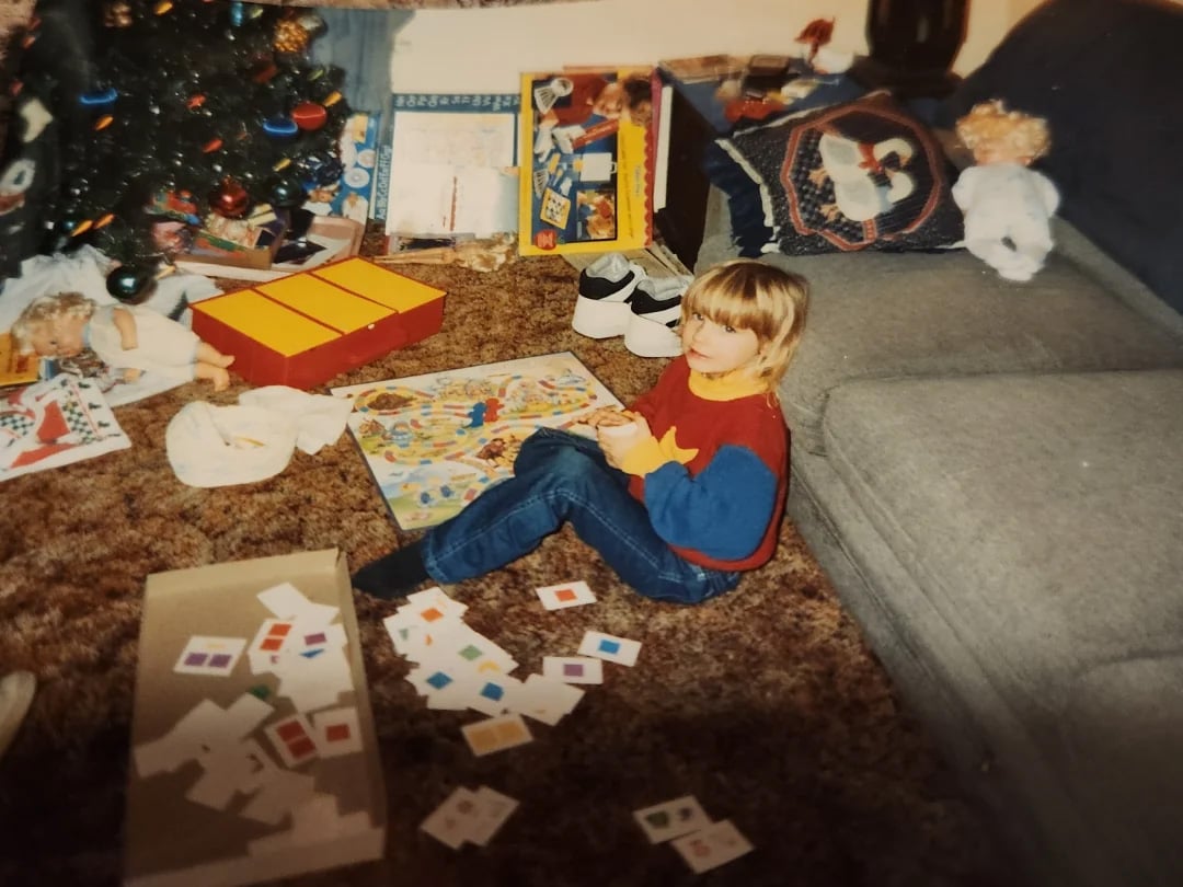 A young child sits on a brown carpet playing a board game near a gray couch and a decorated Christmas tree. Game cards are scattered around, and toys and gifts are visible in the background.