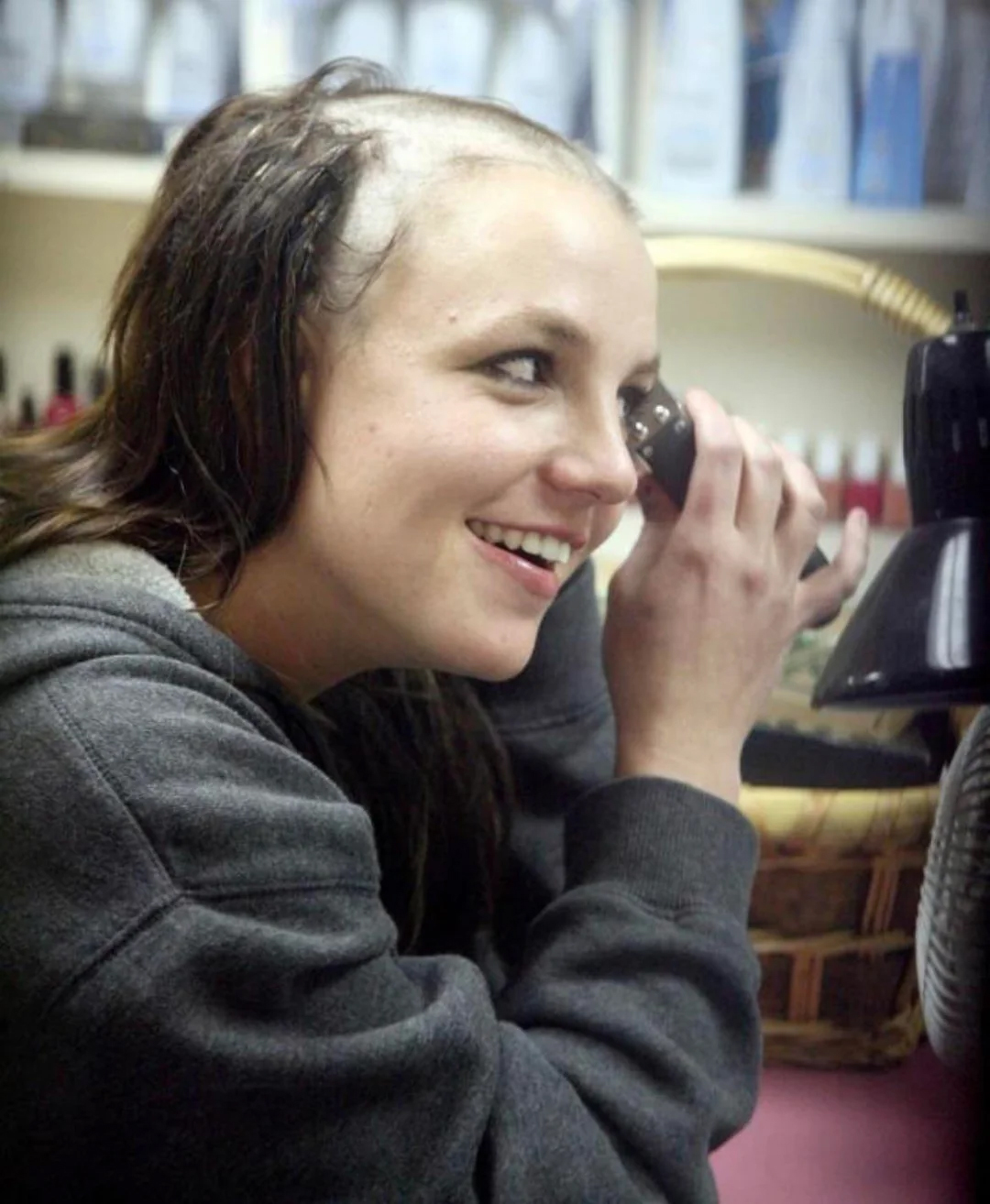 A woman in a gray hoodie smiles while shaving her head with electric clippers, revealing a partially shaved scalp. She is indoors, sitting by a table with baskets and various items in the background.