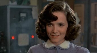 A young woman with wavy brown hair and a light purple sweater with a white collar smiles softly. The background is slightly blurred, showing what appears to be vintage equipment or machinery.