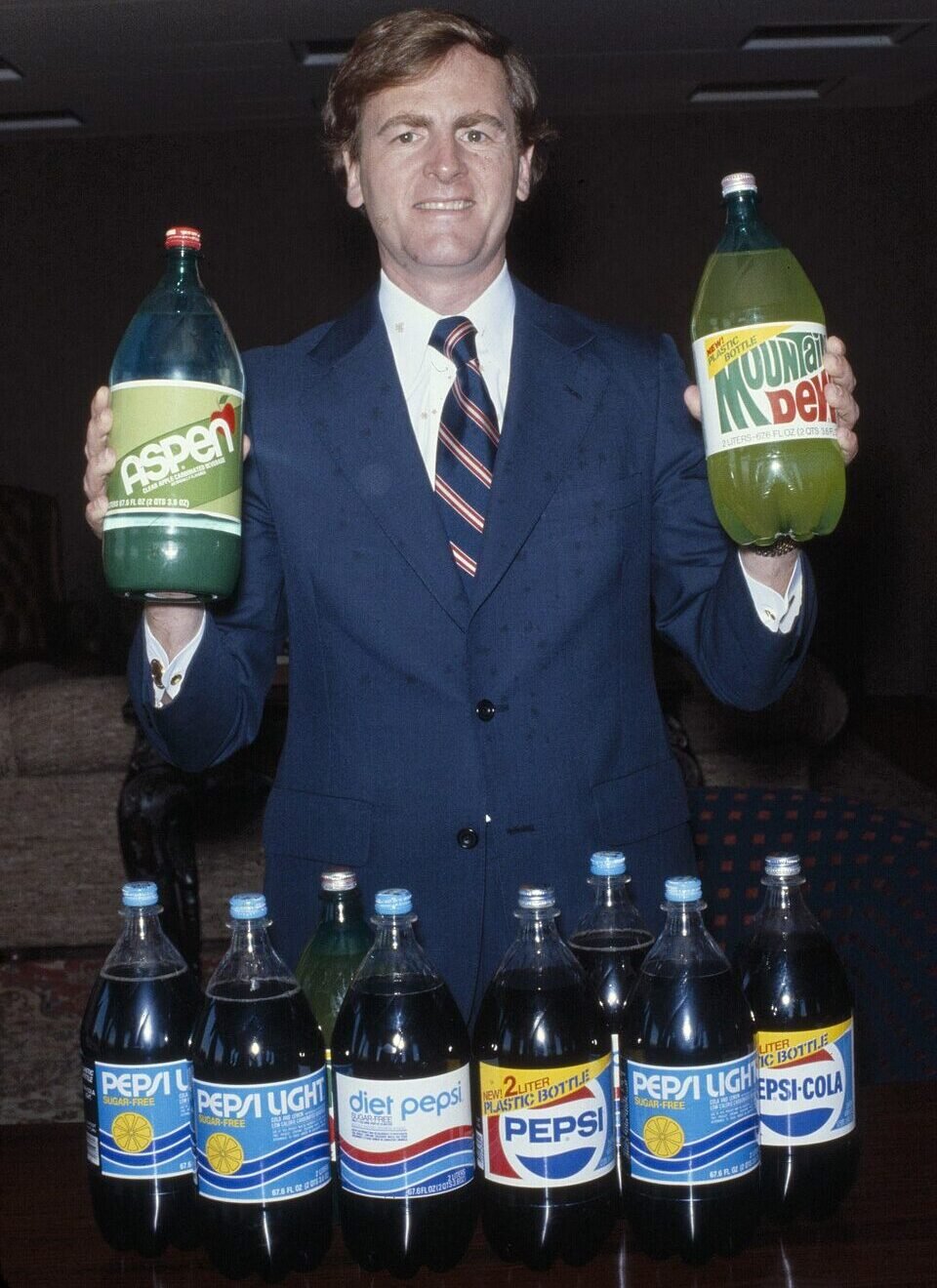 A man in a suit and tie smiles while holding large bottles of Aspen and Mountain Dew soda. In front of him, several bottles of Pepsi, Diet Pepsi, and Pepsi Light are lined up on a table.