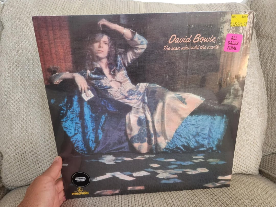 A hand holds a vinyl record of David Bowie's "The Man Who Sold the World," featuring Bowie in a long dress reclining on a couch. Price and sale stickers are on the album's plastic cover.