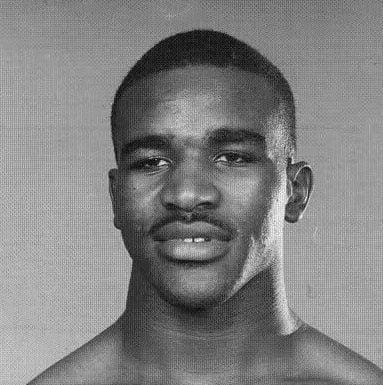 A black and white portrait of a young, shirtless man with short hair, a mustache, and a neutral expression, looking directly at the camera.