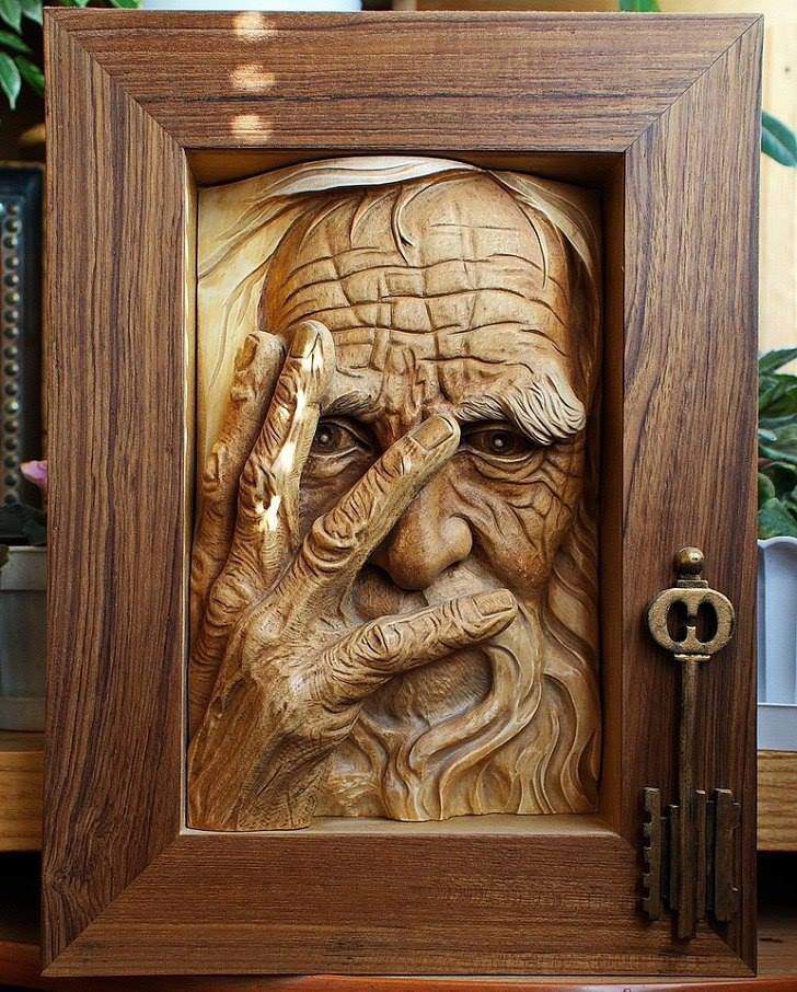 A detailed wooden carving of an elderly man's face and hand, framed in wood. The man has deep wrinkles, a long beard, and his hand partly covers his face. A large, ornate key is attached to the right side of the frame.