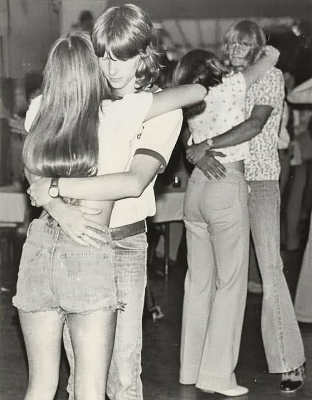 Black and white photo of two young couples slow dancing. The pair in the foreground, a girl in denim shorts and a boy with long hair, embrace closely. Another couple is dancing in the background.