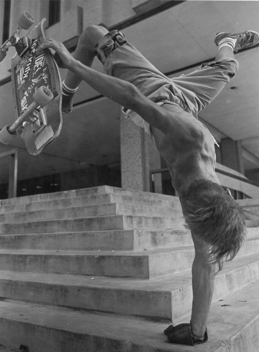 A shirtless skateboarder performs a one-handed handstand on a skateboard at the top of concrete steps, with his legs in the air and his board held by one hand. The background shows a building with columns and railings.