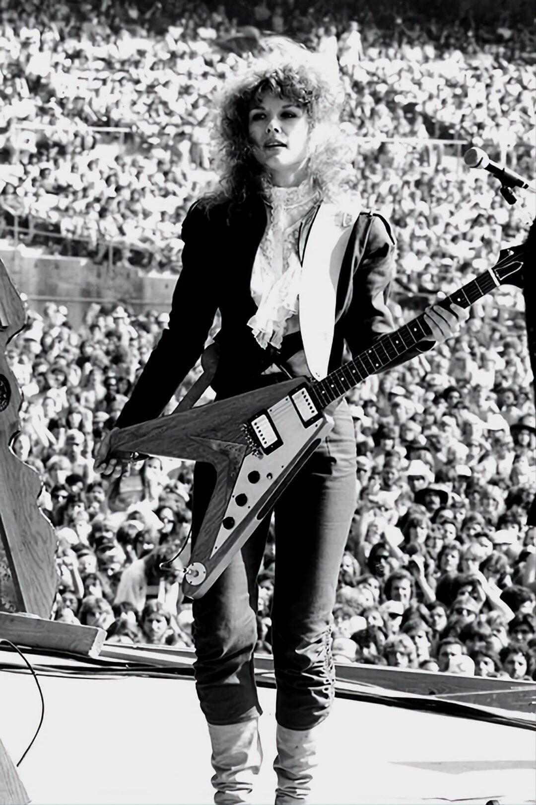 A musician with curly hair in a dark outfit and boots stands on an outdoor stage, holding a Flying V electric guitar, with a large crowd of people in the background. The image is black and white.