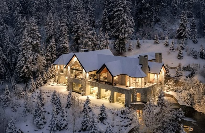 A large, warmly lit stone and wood house stands surrounded by snow-covered trees and hills in a winter forest setting at dusk. Snow blankets the roof and grounds, creating a cozy, serene atmosphere.