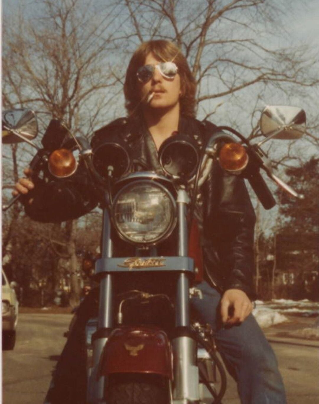 A person wearing sunglasses and a leather jacket sits on a motorcycle, facing the camera. Leafless trees and a car are visible in the background on a sunny day.