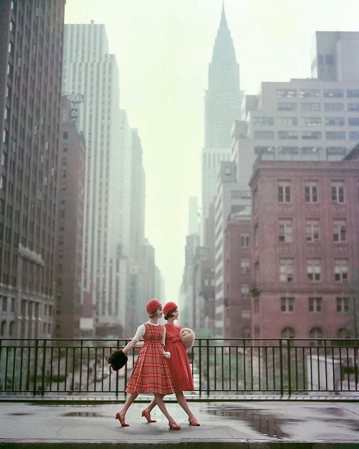 Two women in matching red outfits and hats walk across a city street, carrying handbags, with tall buildings and the Chrysler Building visible in the background on a misty day.