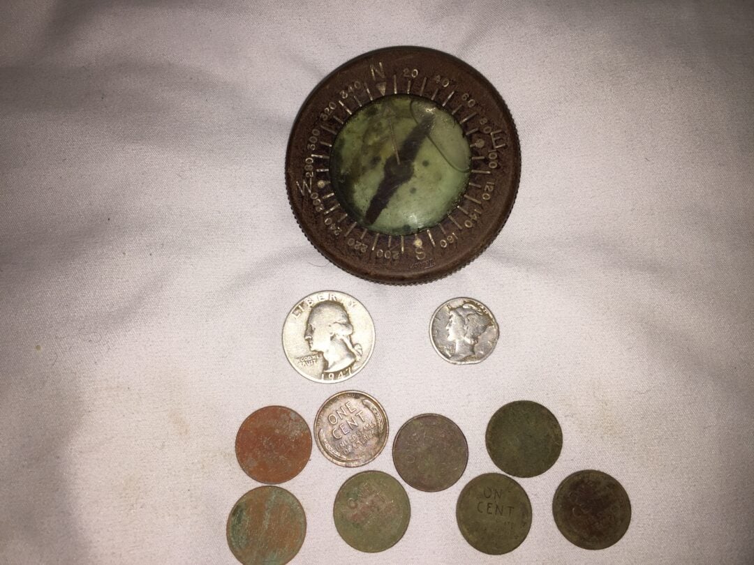 A tarnished compass sits above a group of coins, including a U.S. quarter, a dime, and several worn pennies, all arranged on a white fabric background.