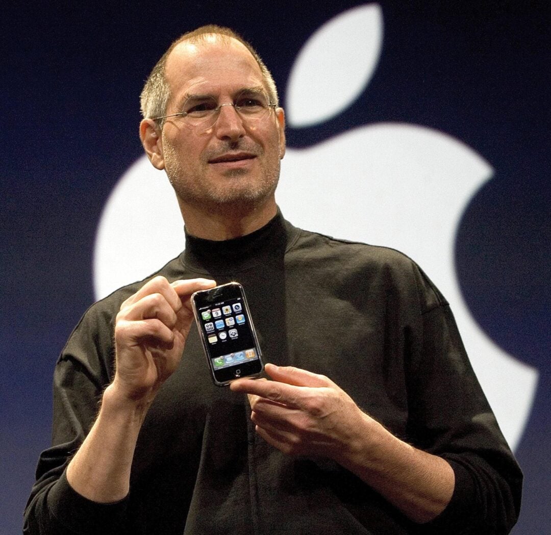 A man in a black turtleneck holds up an early model iPhone in front of a large white Apple logo on a dark background, appearing to present or introduce the device.