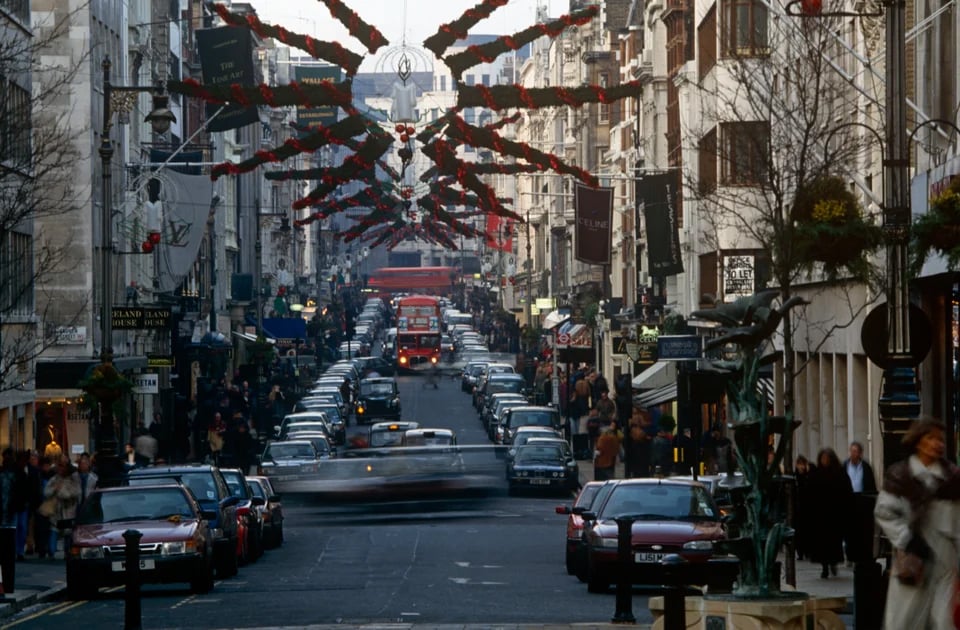 A busy city street decorated with red and green garlands for the holidays, lined with parked cars, shops, and pedestrians, with red double-decker buses in the distance.