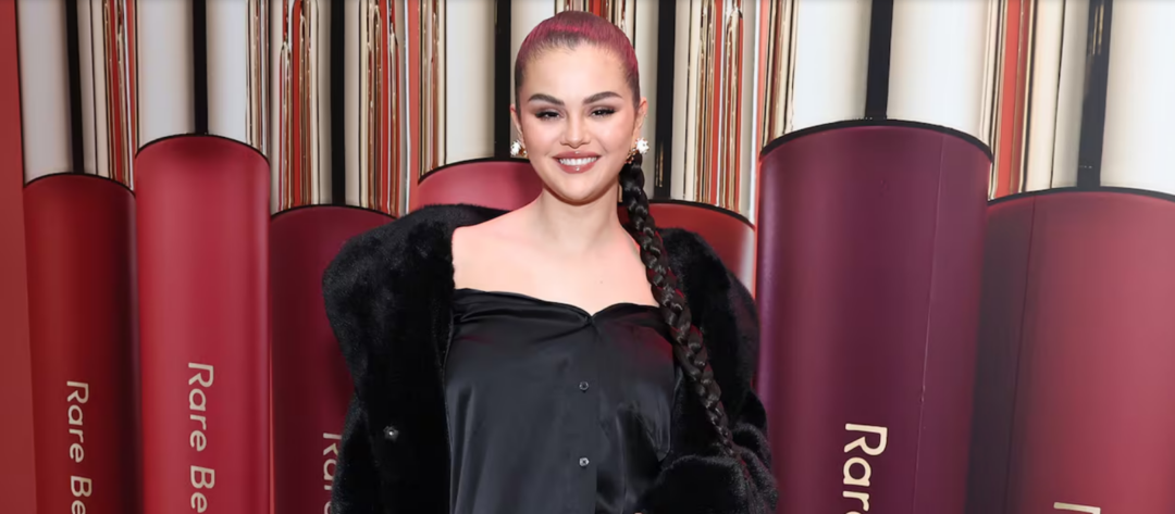 A woman with a long braided ponytail, wearing a black off-the-shoulder outfit and a dark fur coat, stands smiling in front of a backdrop featuring oversized tubes of Rare Beauty lip products.
