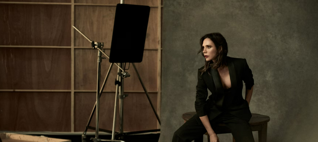 A woman in a black suit sits on a stool in a studio with a gray backdrop. She looks to the side, while studio lighting equipment and a wooden set wall are visible in the background.