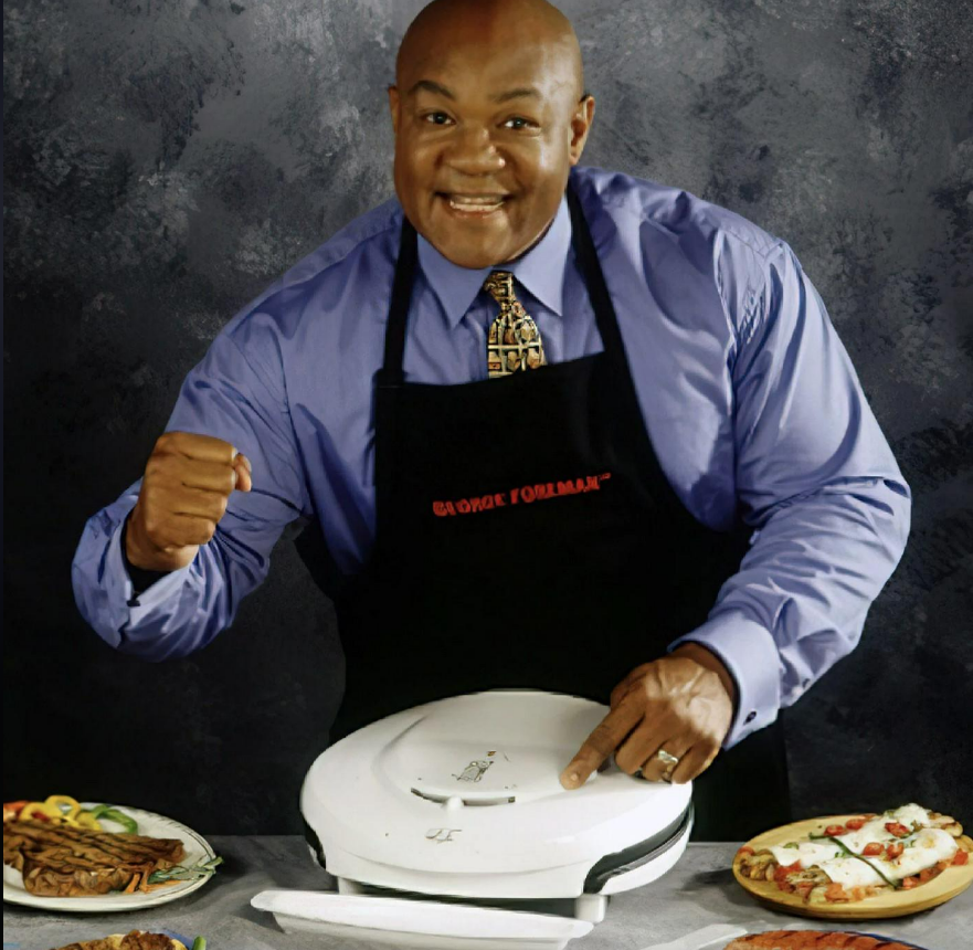 A smiling man wearing a blue shirt, patterned tie, and black apron stands behind a countertop, holding a closed George Foreman Grill with various cooked foods displayed on plates around him.