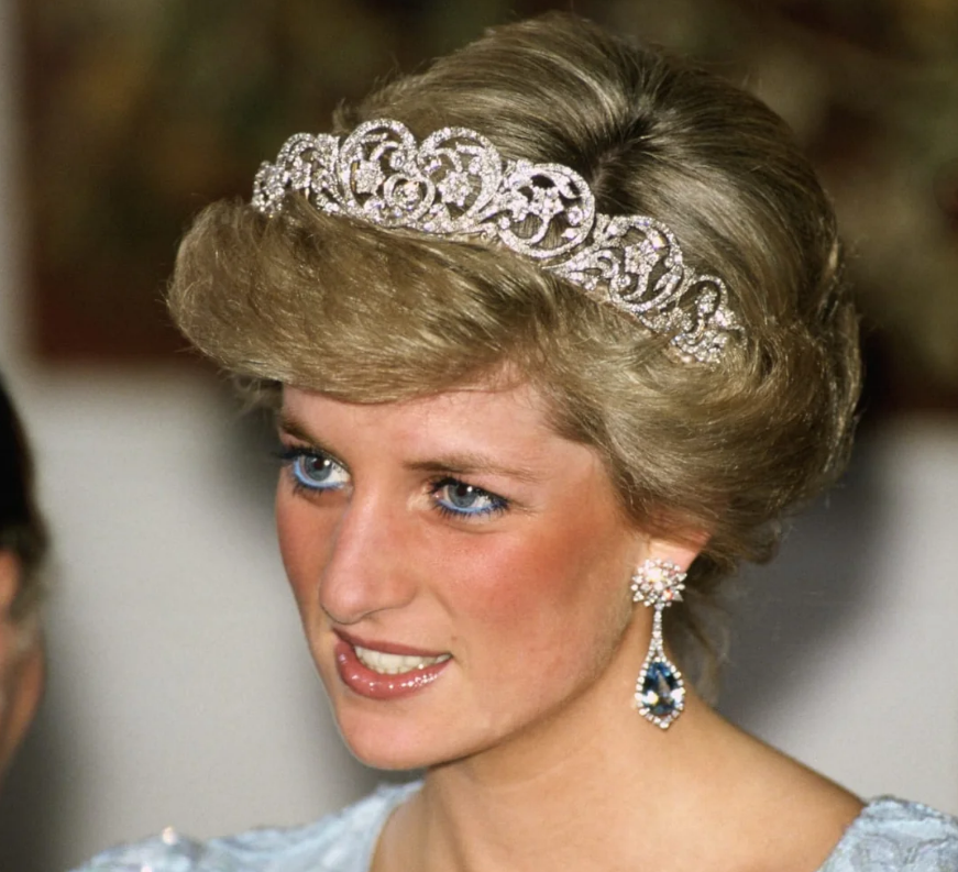 A woman with short blonde hair wears a sparkling diamond tiara, matching blue and silver earrings, and a light blue dress. She has bright blue eyes and a gentle smile.
