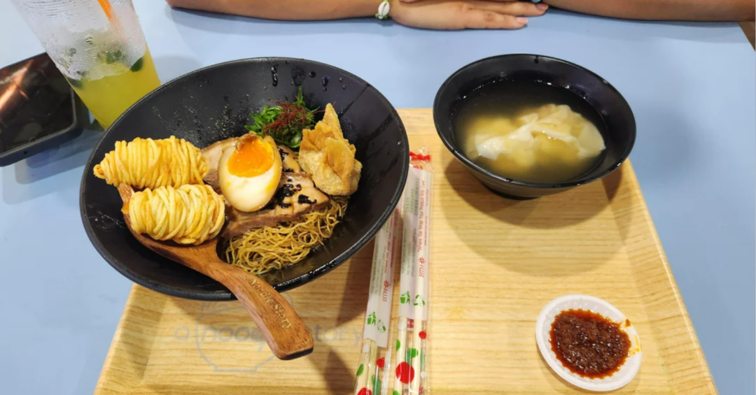 A tray with a bowl of noodles topped with egg, meat, fried items, and greens, a bowl of clear soup with dumplings, a small dish of chili sauce, two pairs of chopsticks, a green drink, and a person's arms resting on the table.