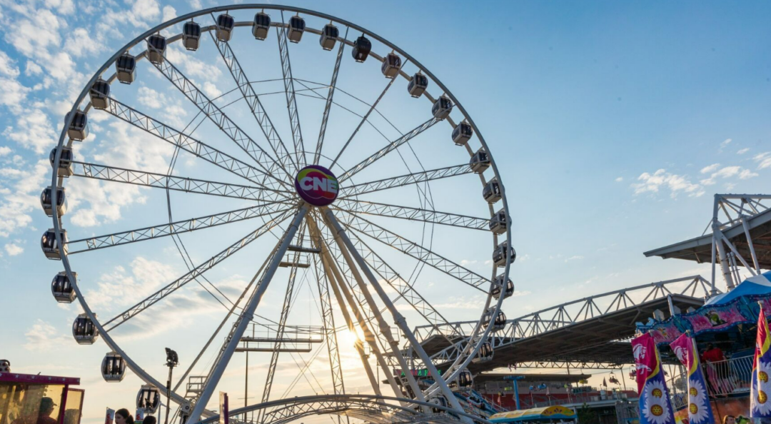 A large Ferris wheel with a "CNE" sign at its center stands against a blue sky at sunset, surrounded by fair booths and structures at a bustling outdoor event.