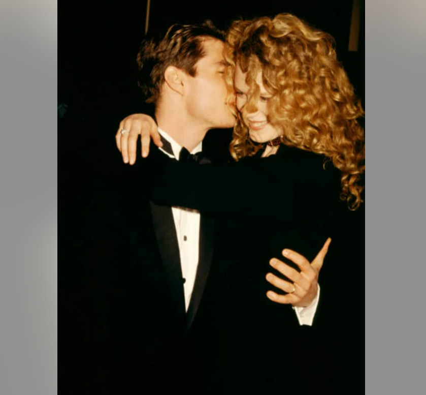 A man in a tuxedo and a woman with long curly hair embrace closely, smiling and touching foreheads in an affectionate, dimly lit setting.