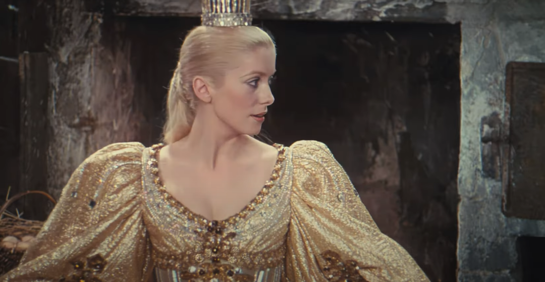 A woman with blonde hair in an elegant gold dress and tall crown looks to her left, standing in front of a rustic, dark stone background.