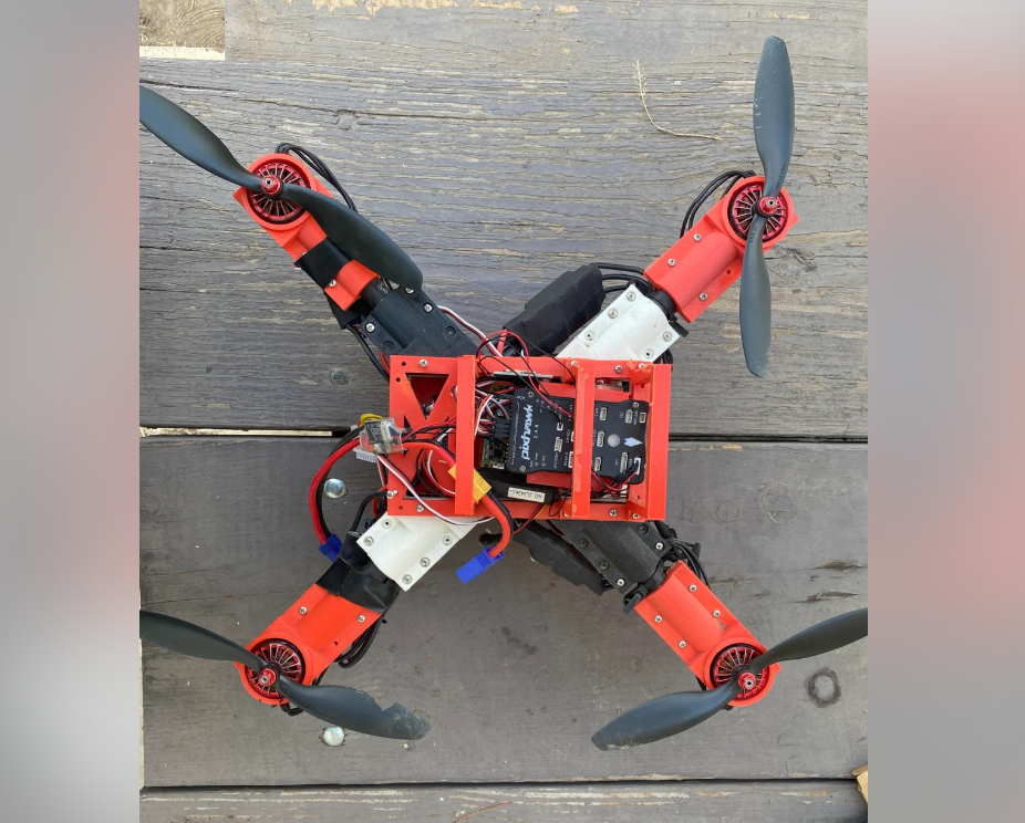 A homemade quadcopter drone with red and black arms, exposed wiring, and visible electronic components rests on a wooden surface. The propellers are positioned at the ends of each arm.