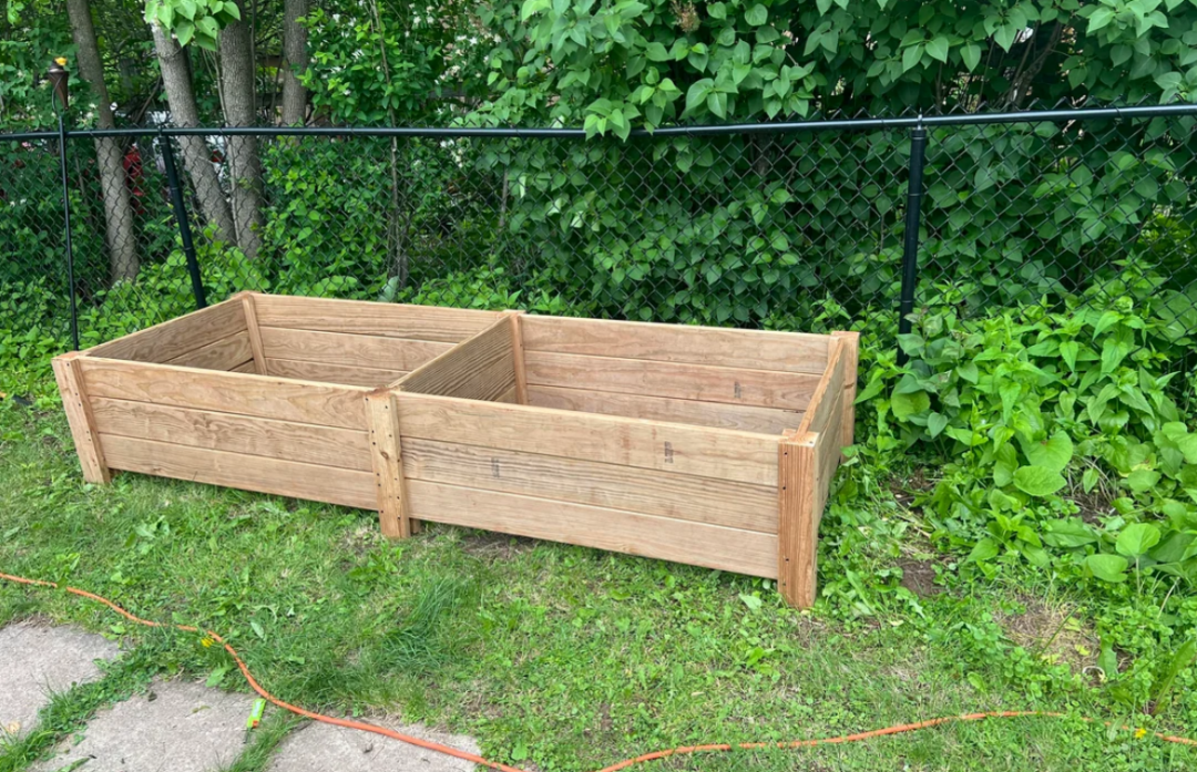 Two empty wooden raised garden beds are placed on a grassy area near a black chain-link fence, with green leafy bushes and trees in the background. An orange extension cord lies on the grass nearby.