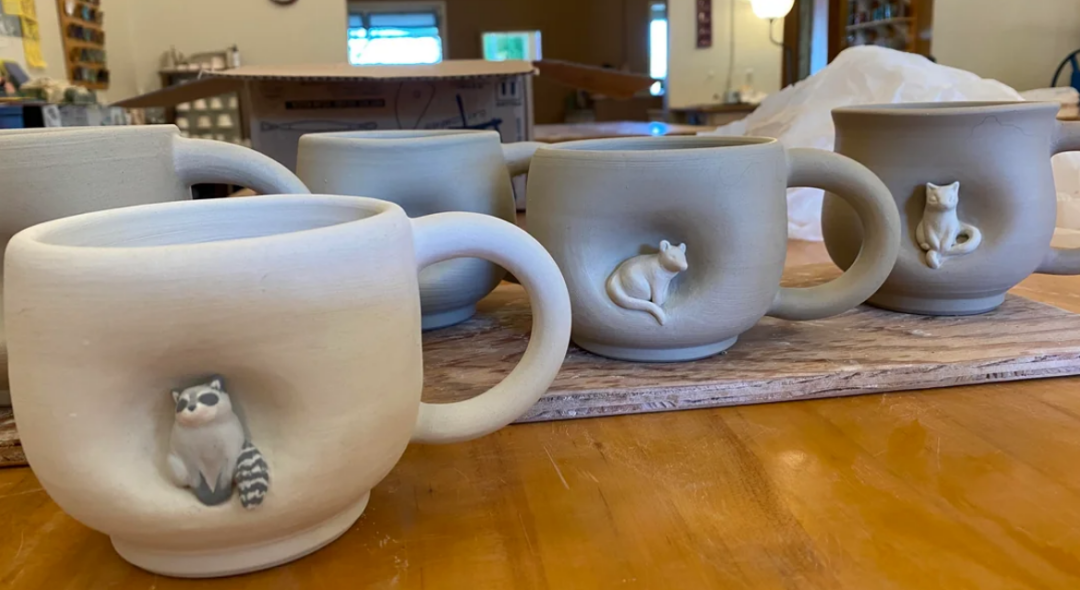 Five unglazed ceramic mugs with animal figures, including a raccoon, mouse, and cat sculpted on the sides, sit on a wooden board on a table in a pottery studio with shelves and warm lighting in the background.