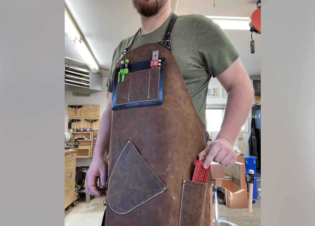 A person in a green shirt wears a leather apron with pens and small tools in the chest pocket, standing in a workshop and placing a red tool into a front pocket. Shelves and equipment are visible in the background.