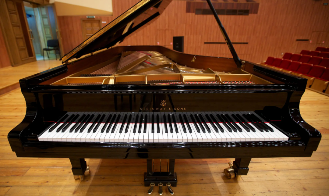 A black Steinway & Sons grand piano with its lid open, positioned on a wooden stage in an empty concert hall with red seats in the background.