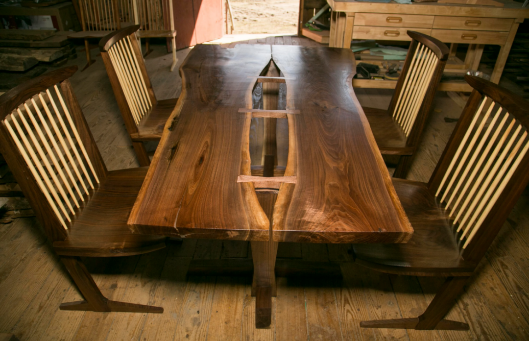 A handcrafted wooden dining table with a natural live edge and butterfly joints, surrounded by four matching wooden chairs with vertical slats, set on a wooden floor in a workshop.