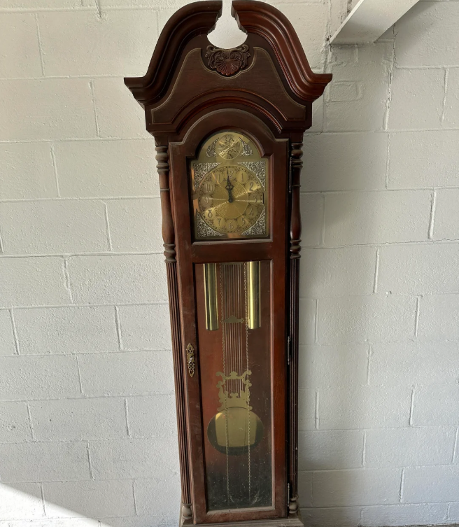 A tall antique wooden grandfather clock with ornate details, a glass front, gold clock face, and visible pendulum stands against a white cinder block wall.