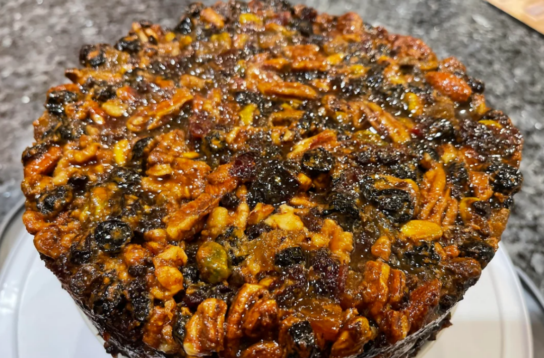 A close-up of a dense fruitcake topped with glazed nuts and dried fruits, including pecans, pistachios, and raisins, sitting on a white surface with a blurred dark background.