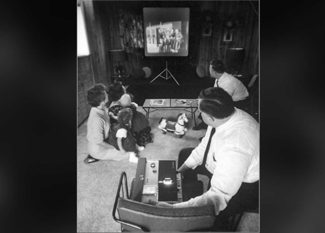 A black-and-white photo shows a family sitting on the floor watching a film projected onto a screen, while a man operates a projector in a living room with wood-paneled walls.