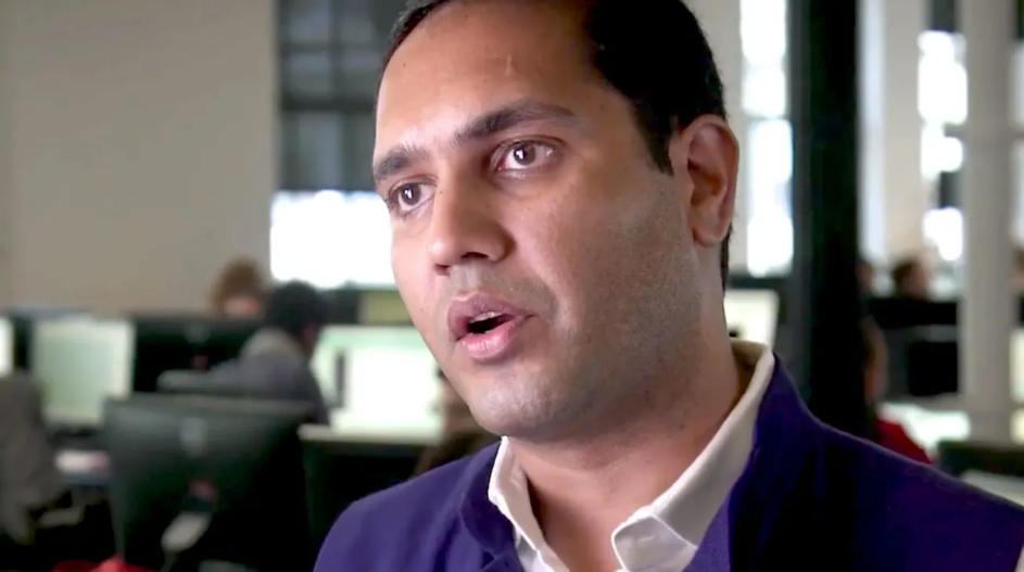 A man in a suit jacket and white shirt speaks in an office setting with blurred people and computer screens in the background.