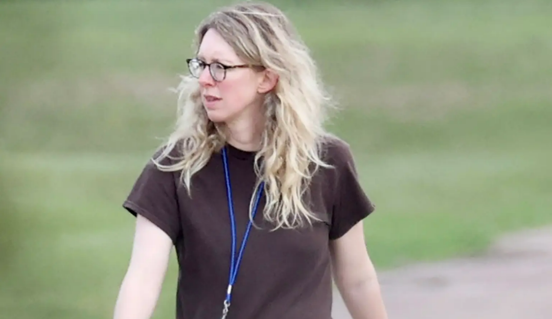 A woman with long blonde hair and glasses, wearing a brown t-shirt and a blue lanyard, stands outdoors on a blurred background of grass and pavement.