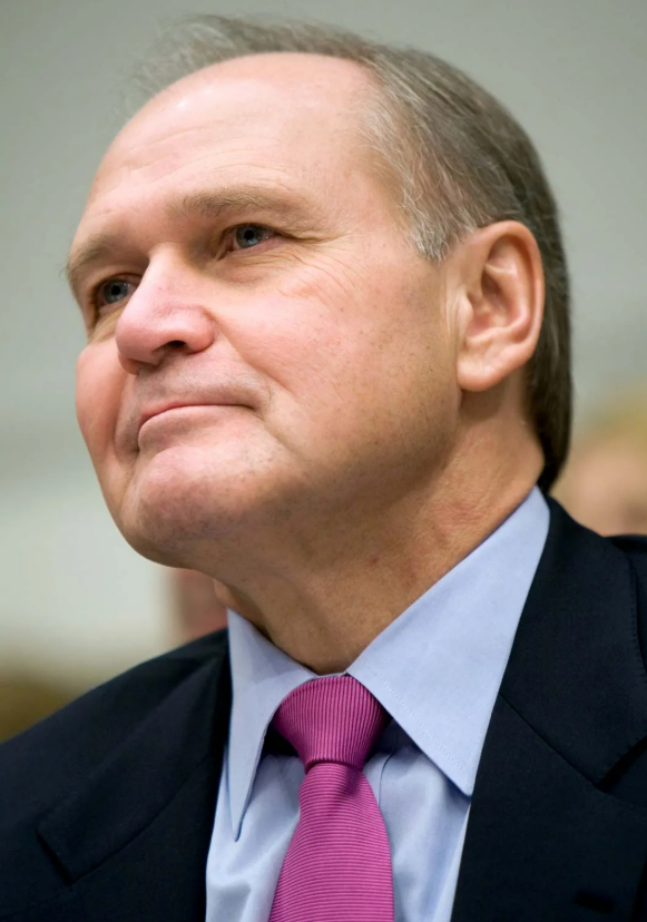 A middle-aged man with short, light brown hair wears a dark suit, light blue shirt, and bright pink tie, looking upward with a neutral expression against a blurred background.