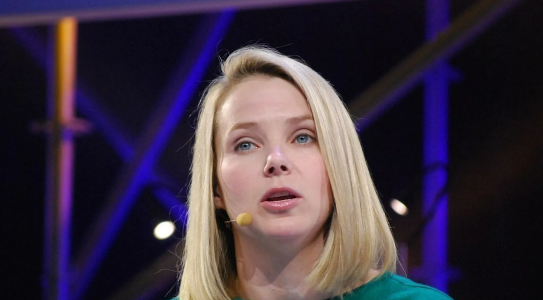A woman with straight blonde hair, wearing a green top and a headset microphone, speaks on stage against a blurred background with blue and purple lighting and metal structures.